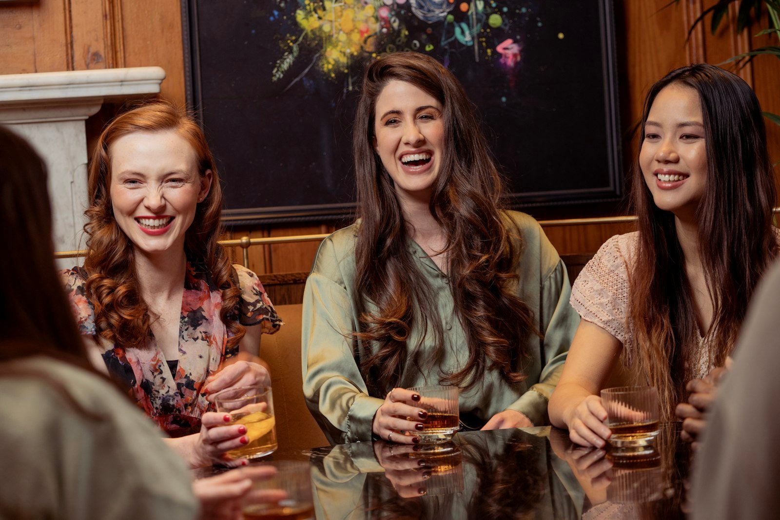 Four women laughing together over drinks at a bar table.